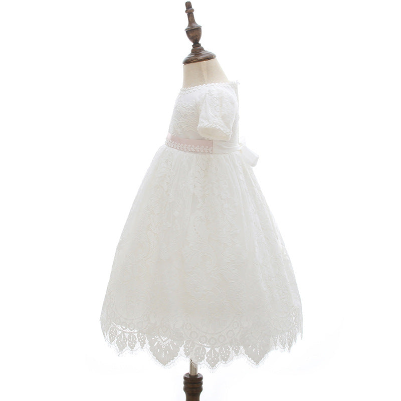Side profile of a baby girl in a white floral lace baptism dress and full matching bonnet, leaning against a neutral armchair at Trendy Angels Boutique, 533 Sydney Rd Brunswick Melbourne.
