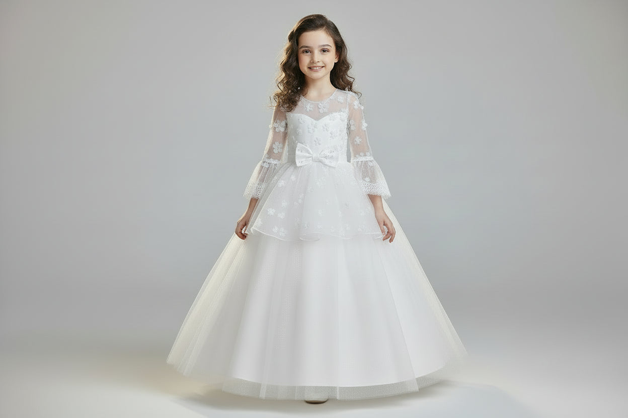 A young girl wearing a floor-length white formal communion dress with long floral lace wing sleeves, a tiered tulle skirt, and a center waist bow at Trendy Angels Boutique.