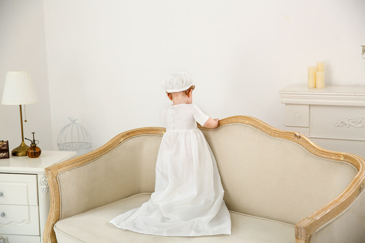 Rear view of a baby boy in a long white heirloom baptism gown and matching bonnet, standing on a beige vintage sofa in a bright room.