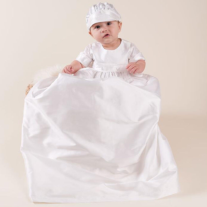 A baby boy wearing a classic white baptism gown and matching cap set sitting in a wicker basket, showcasing high-quality ceremony wear at Trendy Angels.