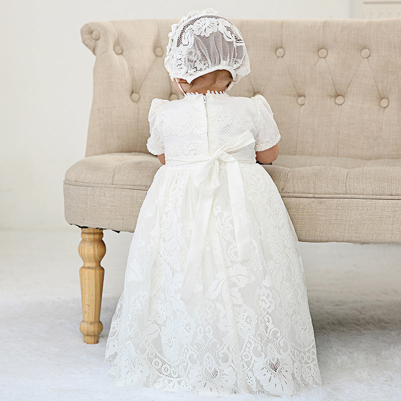 A young child wearing a traditional white lace baptism dress and matching bonnet, showing the elegant back satin bow while sitting on a beige vintage-style couch.