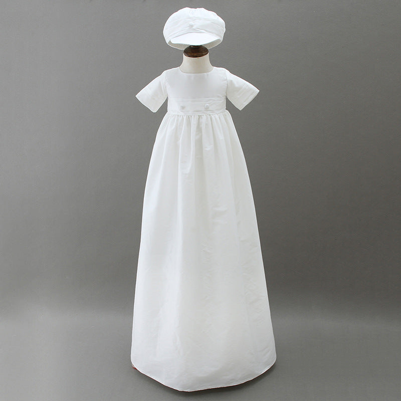 A full-length traditional white baptism gown with a matching hat displayed on a mannequin against a grey background at Trendy Angels Boutique.