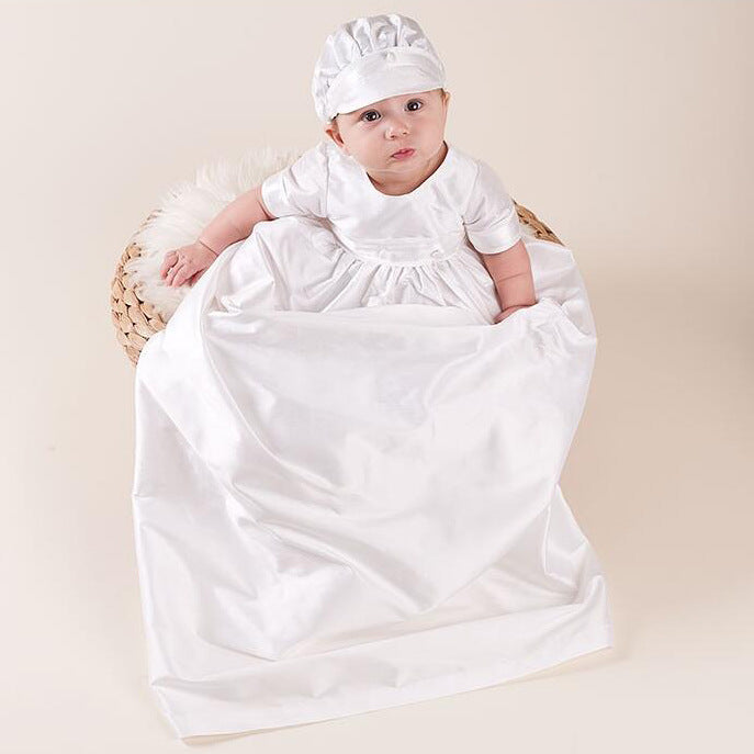 A baby boy wearing a classic white baptism gown and cap set, sitting in a wicker basket to showcase the high-quality fabric.